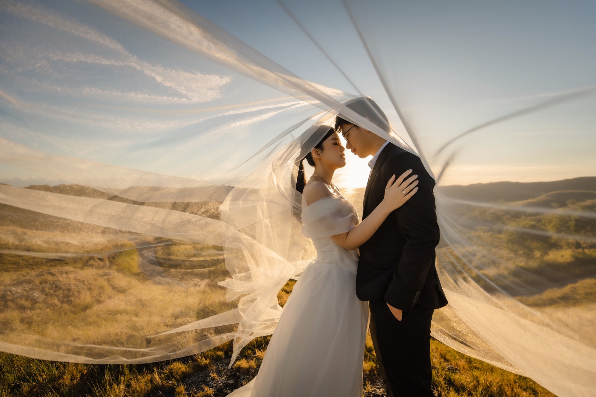 Rey and Feli standing close together beneath a flowing veil in the hills