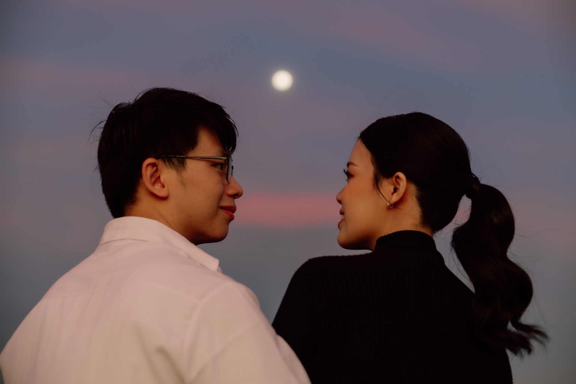 Rey and Feli looking at each other beneath the moon and a pink dusk sky