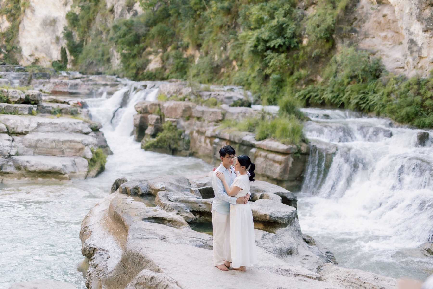 Rey and Feli embracing beside a bright waterfall in a rocky gorge