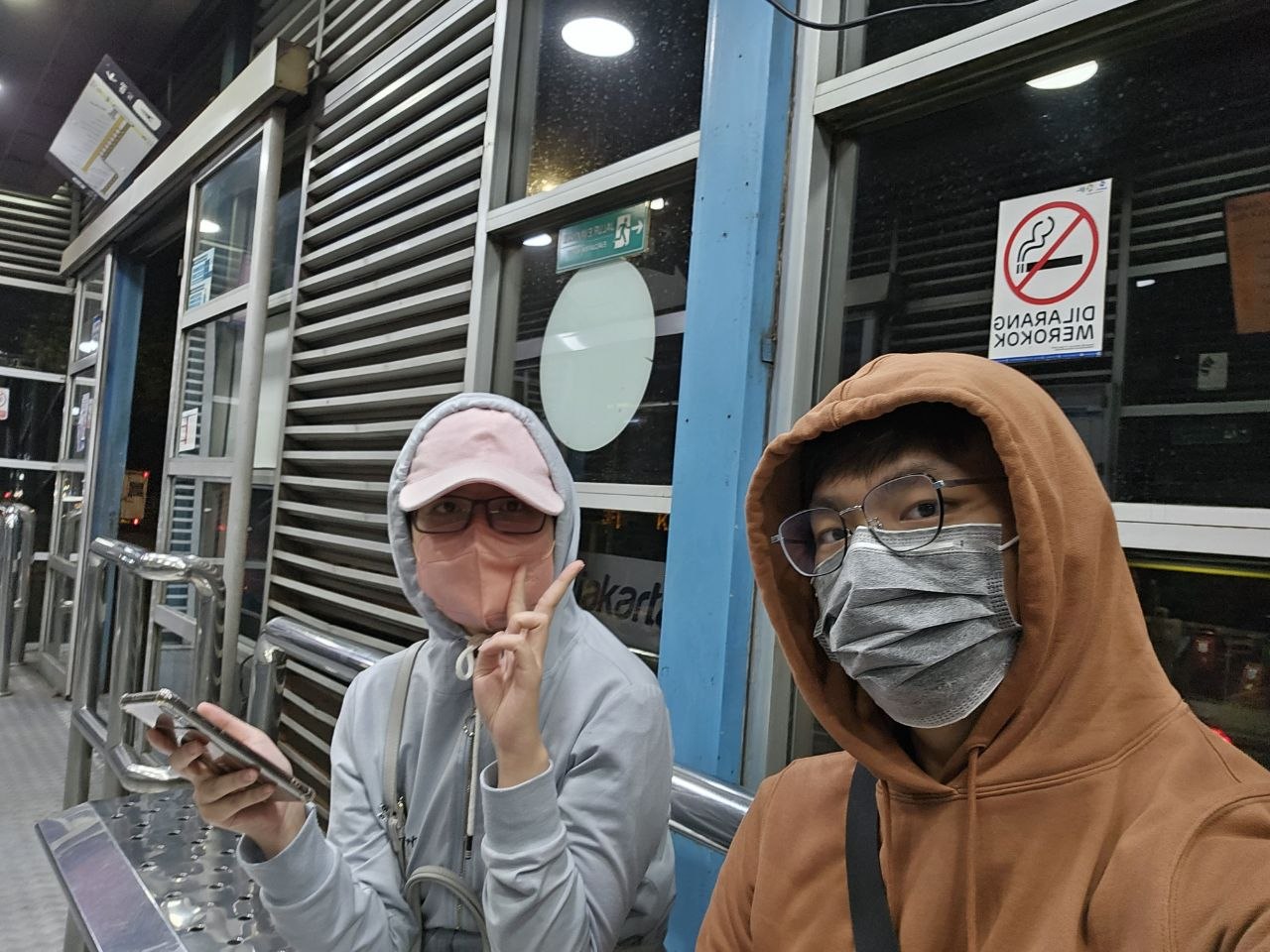 Rey and Feli in hoodies and masks while waiting at a transit stop